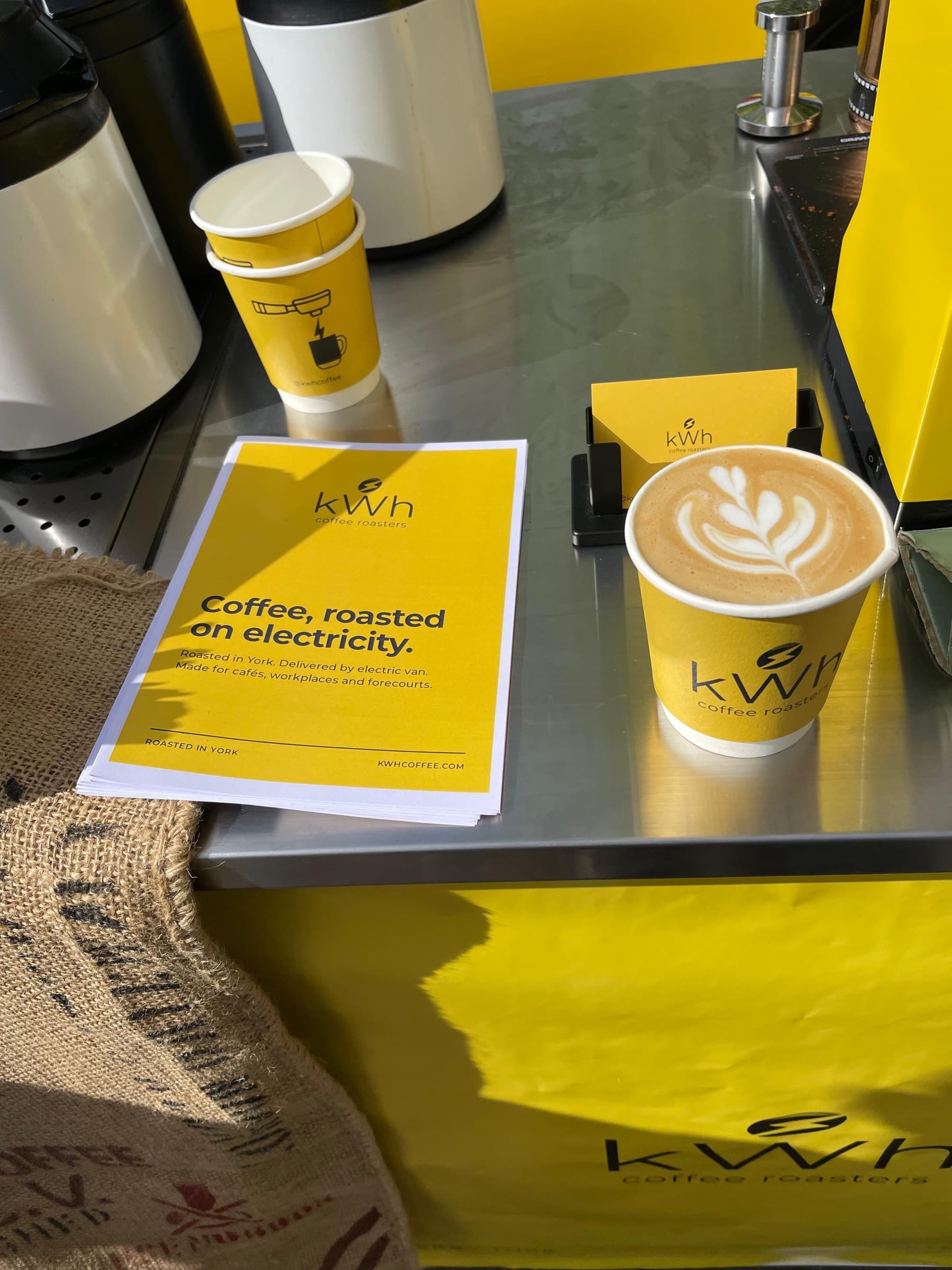 A kWh Coffee flat white with latte art on the bar, beside a leaflet reading Coffee, roasted on electricity