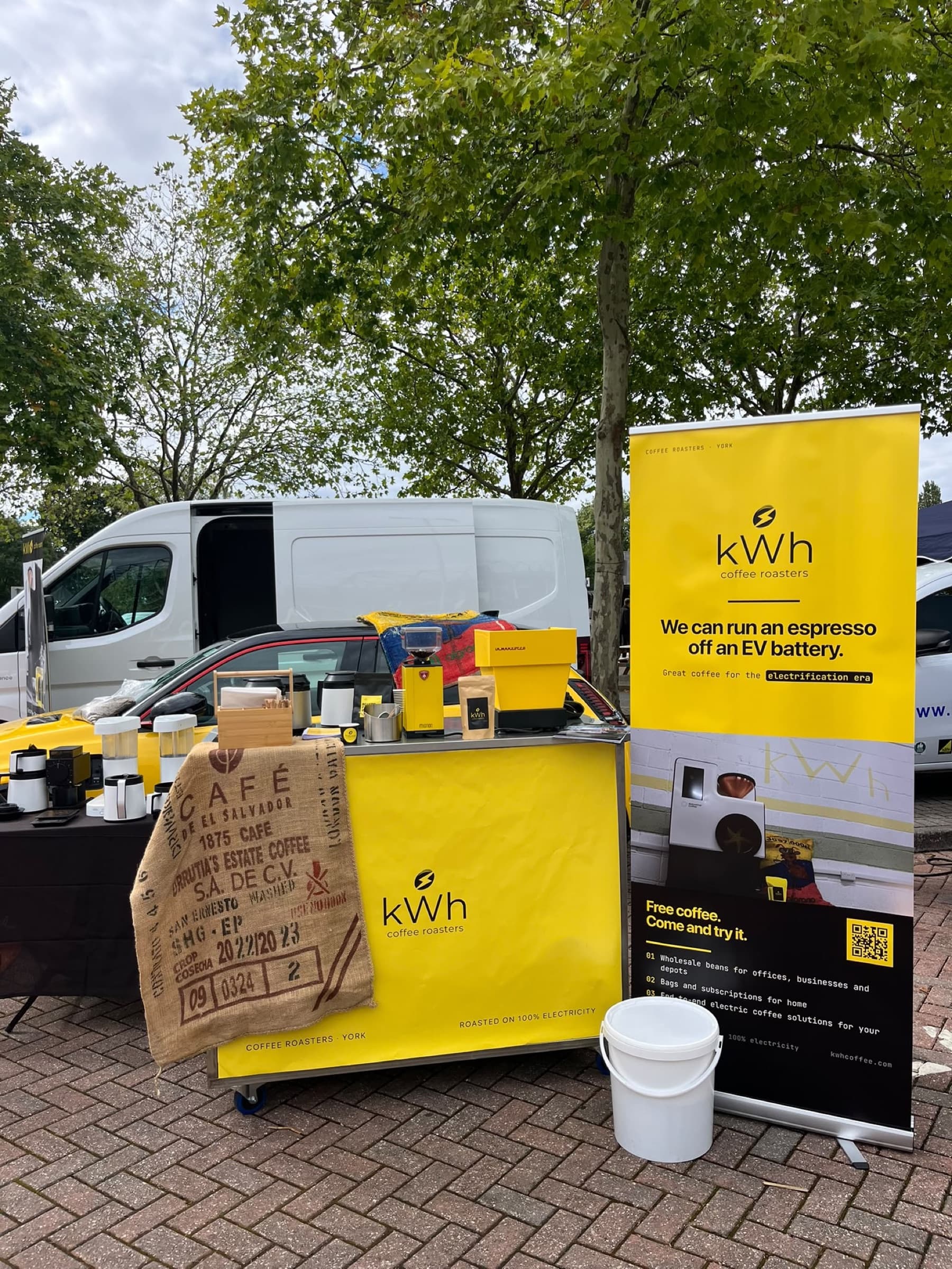 The kWh Coffee Roasters stall at an outdoor event: a yellow coffee cart with a grinder and a coffee sack draped over the front, beside a banner reading 'We can run an espresso off an EV battery'