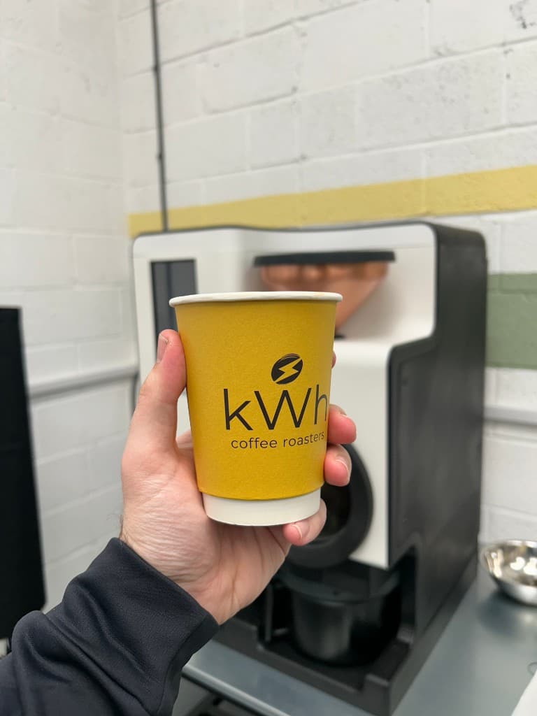 A kWh Coffee Roasters branded cup held in front of the Bellwether electric roaster