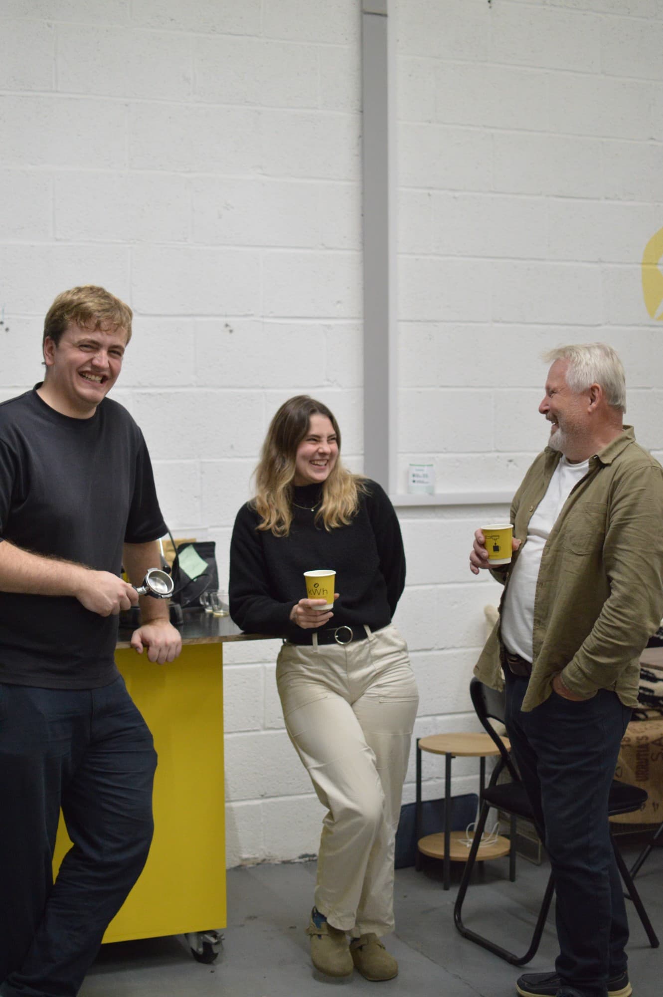 Shaun, Caitlin and Laurence laughing over coffee beside the yellow espresso bar at the roastery